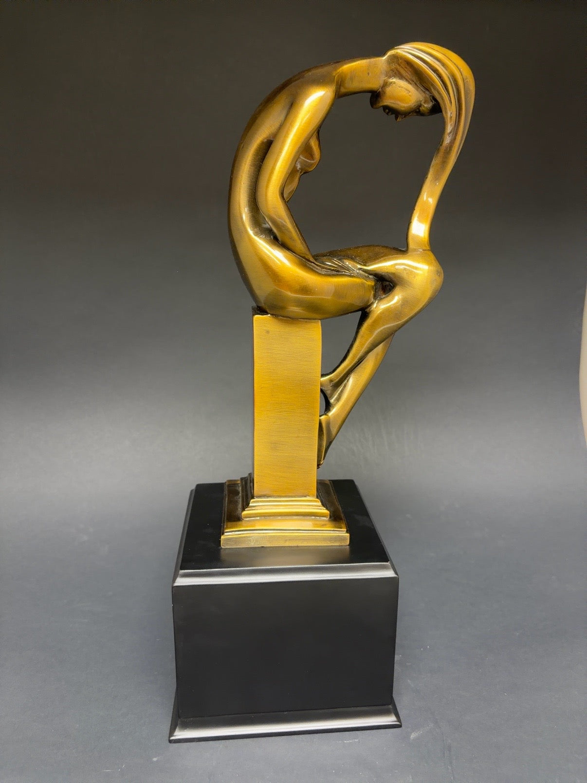 Gold sculpture of a nude figure on a black base against a gray background