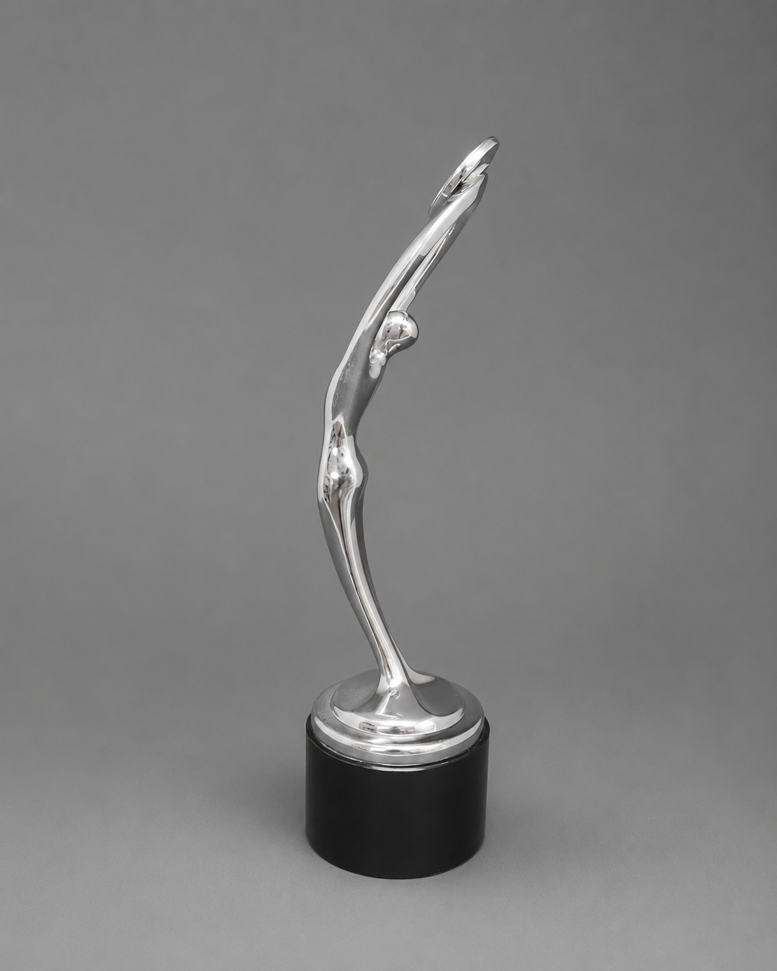 A modern sculptural trophy or award statue with a polished silver abstract figure mounted on a black cylindrical base, photographed against a neutral gray studio background.