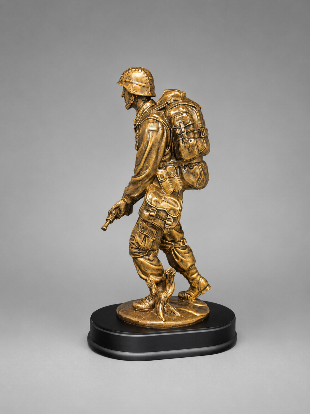 Side view of a bronze-finish military soldier figurine with helmet, backpack, tactical gear, and rifle on a black oval base.
