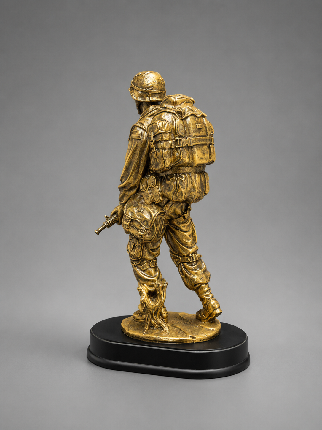 Rear three-quarter view of a detailed bronze-finish soldier figurine showing the backpack, equipment pouches, helmet, and black display base.