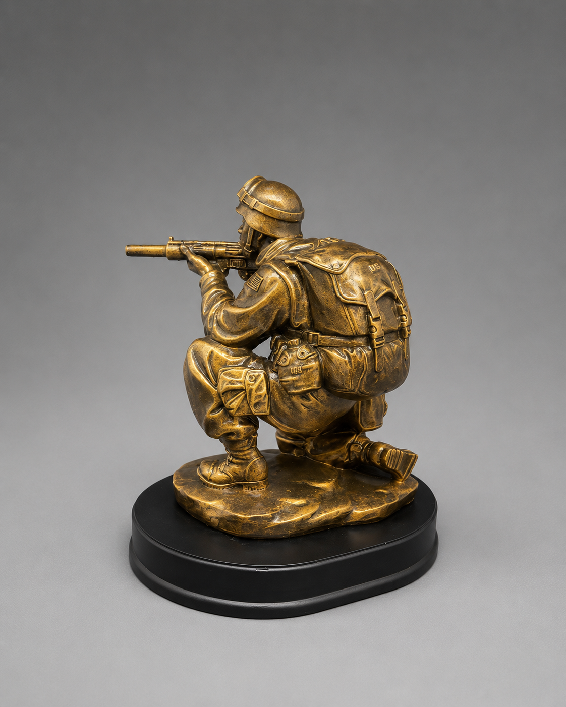 Rear three-quarter view of a bronze-finish kneeling soldier figurine aiming a rifle, showing the detailed backpack, uniform, and black oval base.