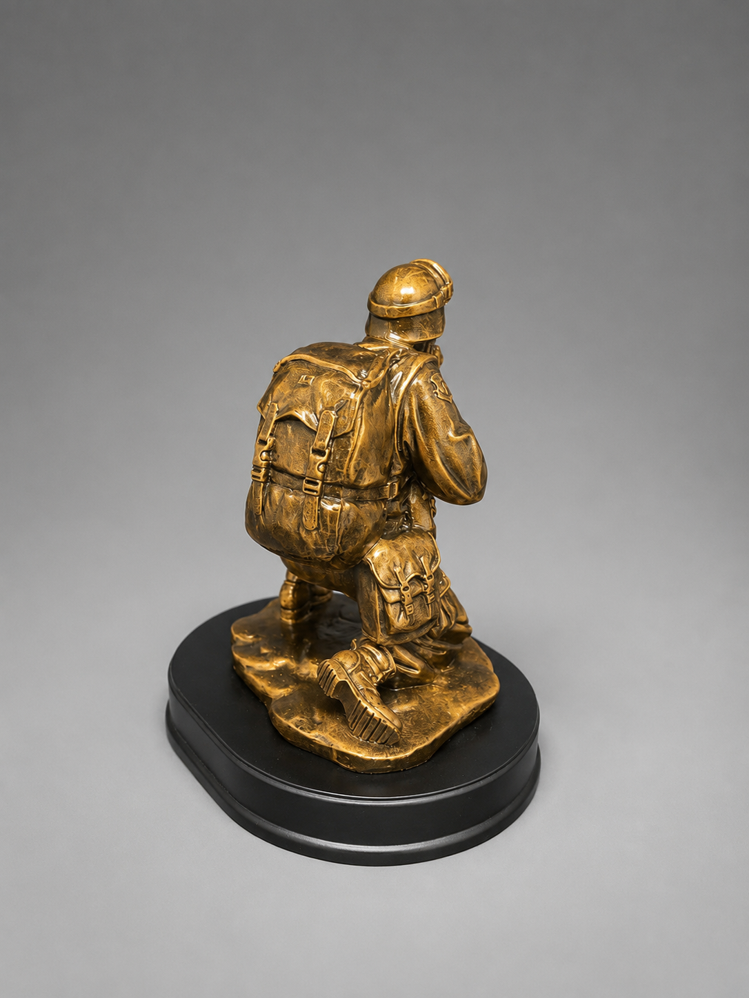 Back view of a bronze-finish kneeling soldier figurine with a detailed backpack and military gear on a black oval display base.