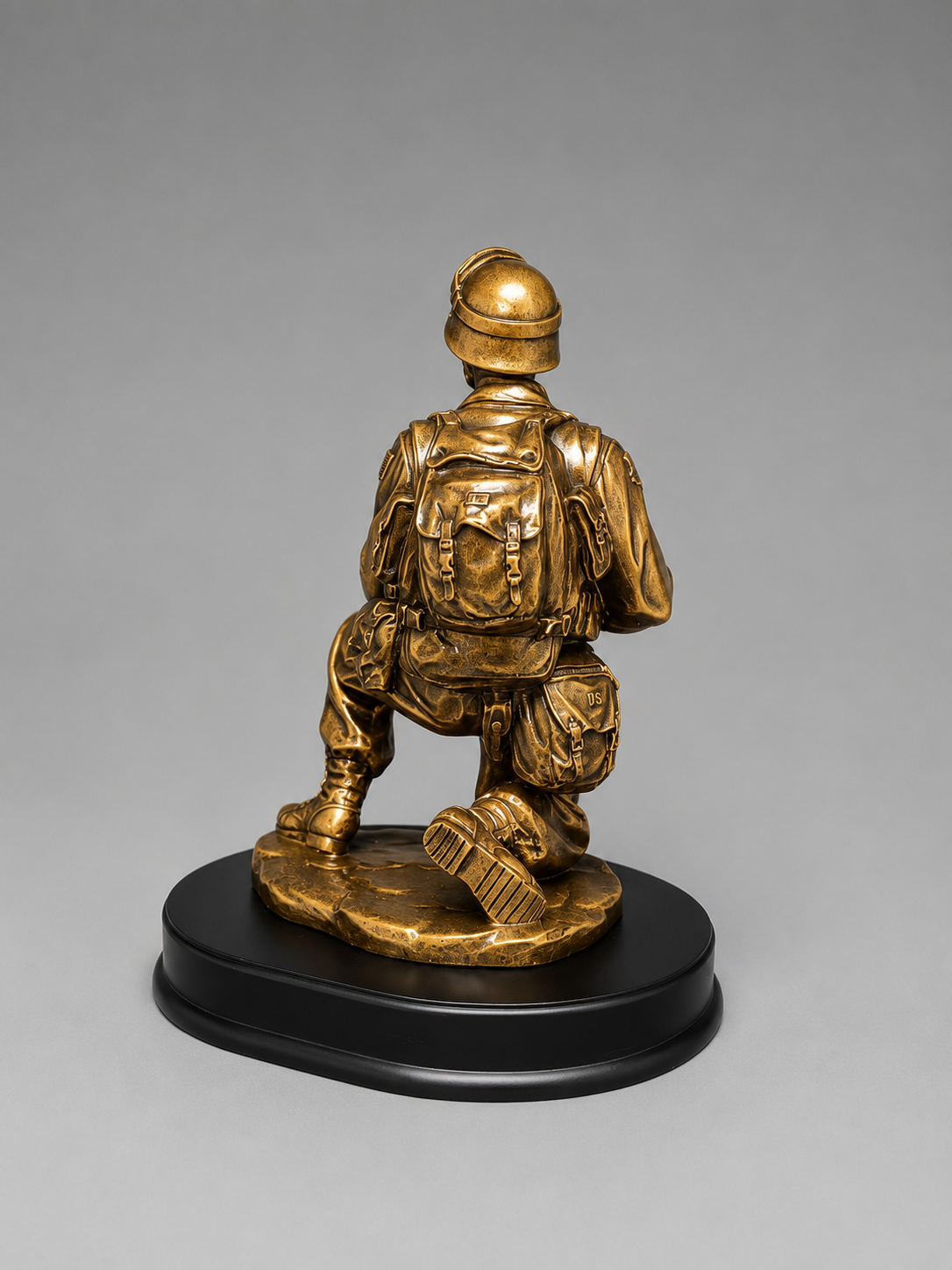 Rear view of an antique gold kneeling soldier figurine showing the detailed helmet, backpack, gear pouches, and black oval base.
