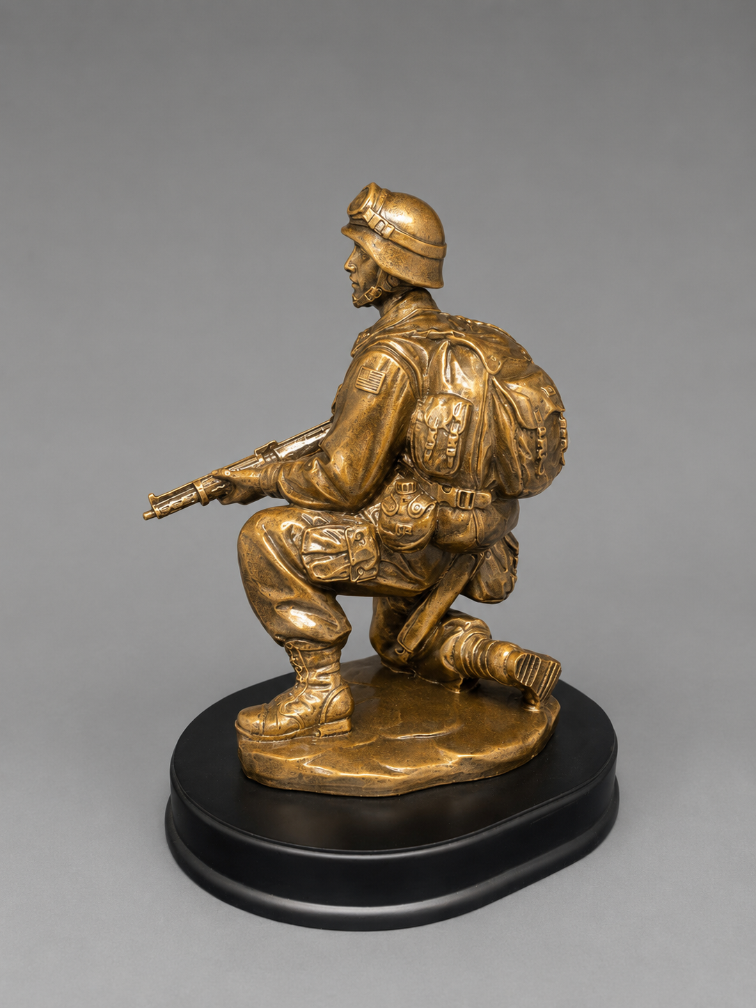 Side and rear angle of an antique gold kneeling soldier figurine with rifle, military backpack, tactical gear, and black display base.