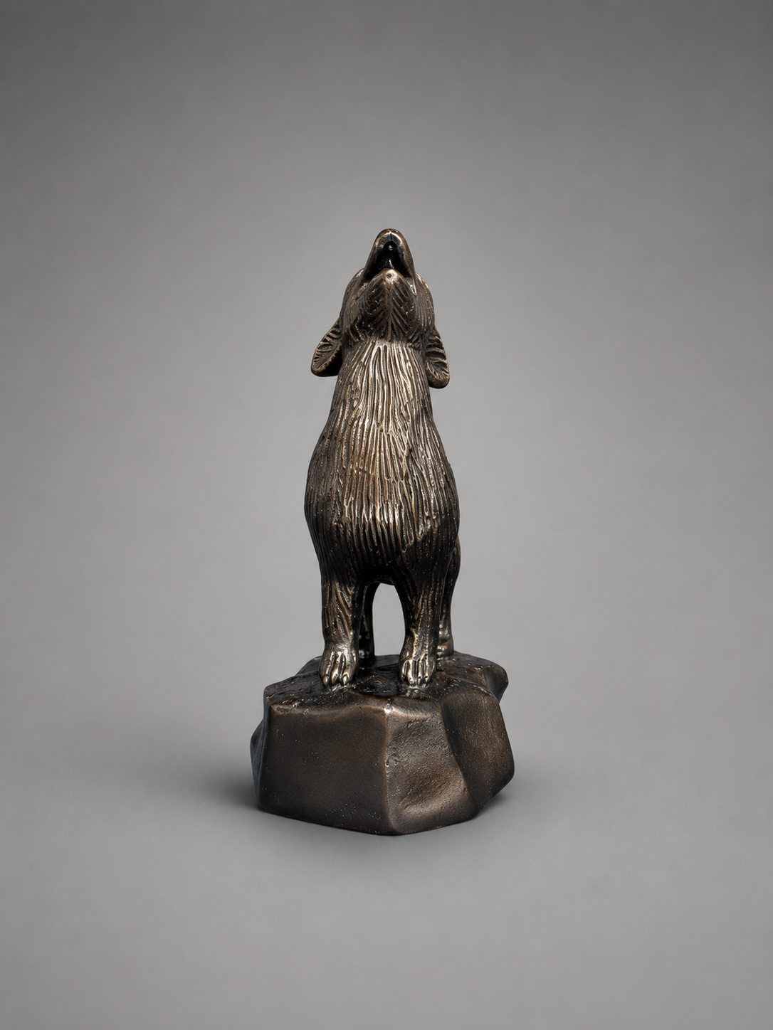 Front view of a metallic howling coyote sculpture standing on a raised rock-style base against a studio gray background.