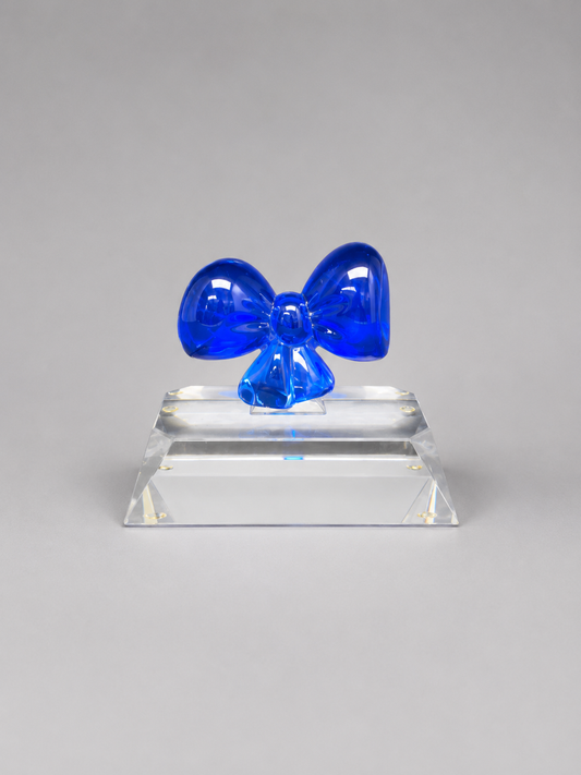 Front view of a blue crystal bow mounted on a clear beveled rectangular base against a neutral gray background.