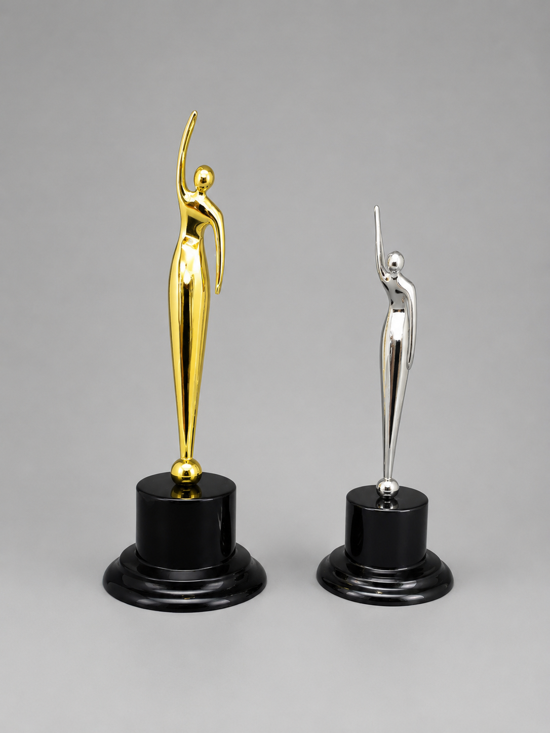 Angled view of modern gold and silver abstract trophy awards with raised-arm figures on black pedestal bases against a gray background.