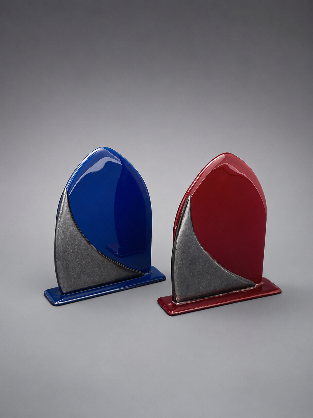 Two glossy geometric bookends with curved, shield-like tops and rectangular bases, shown side by side on a smooth gray studio background. One is blue and silver, and the other is red and silver.