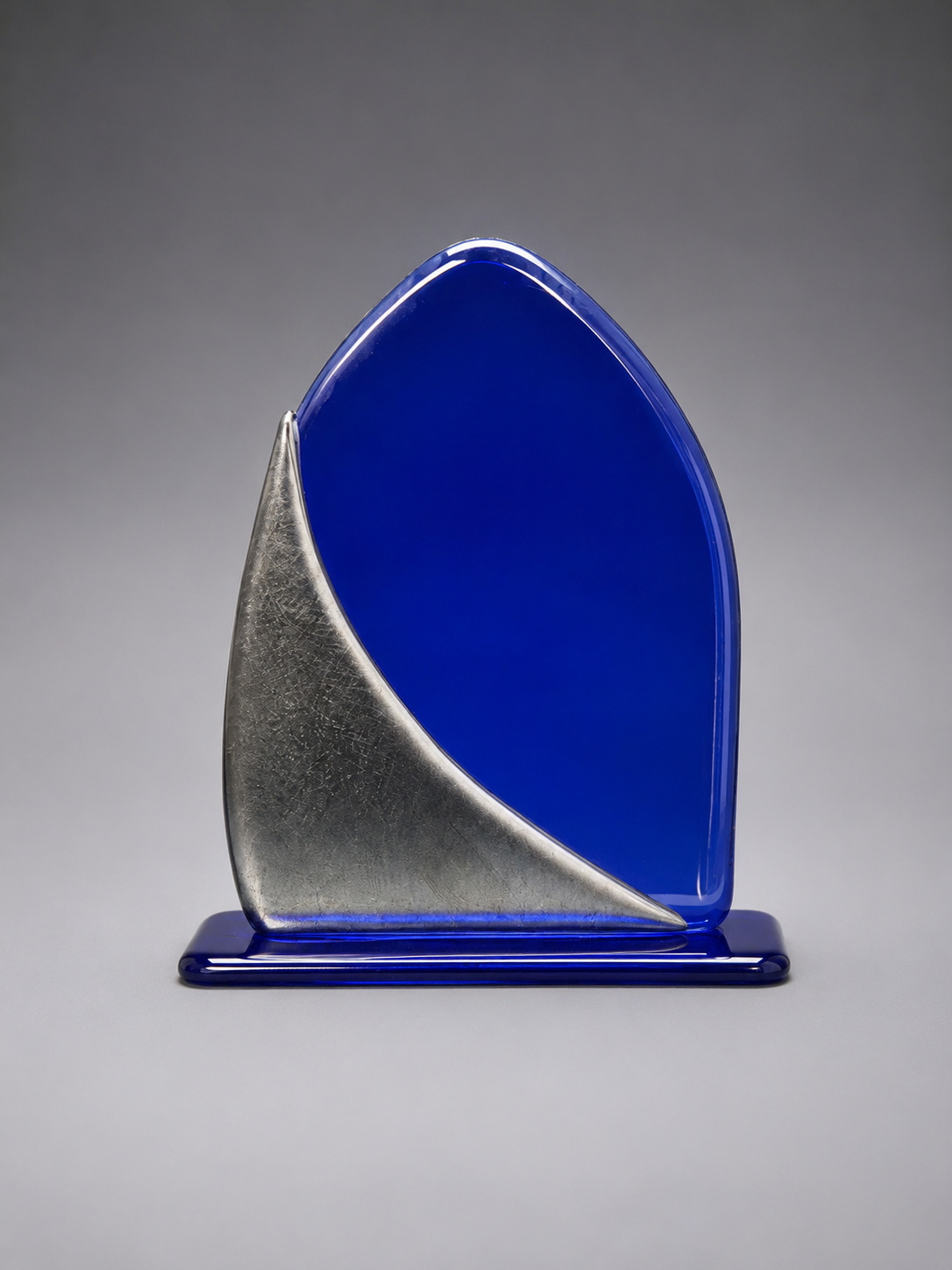 A modern abstract glass award plaque featuring a vibrant cobalt blue color with a textured metallic silver wave design, mounted on a matching blue base against a neutral gray gradient background.