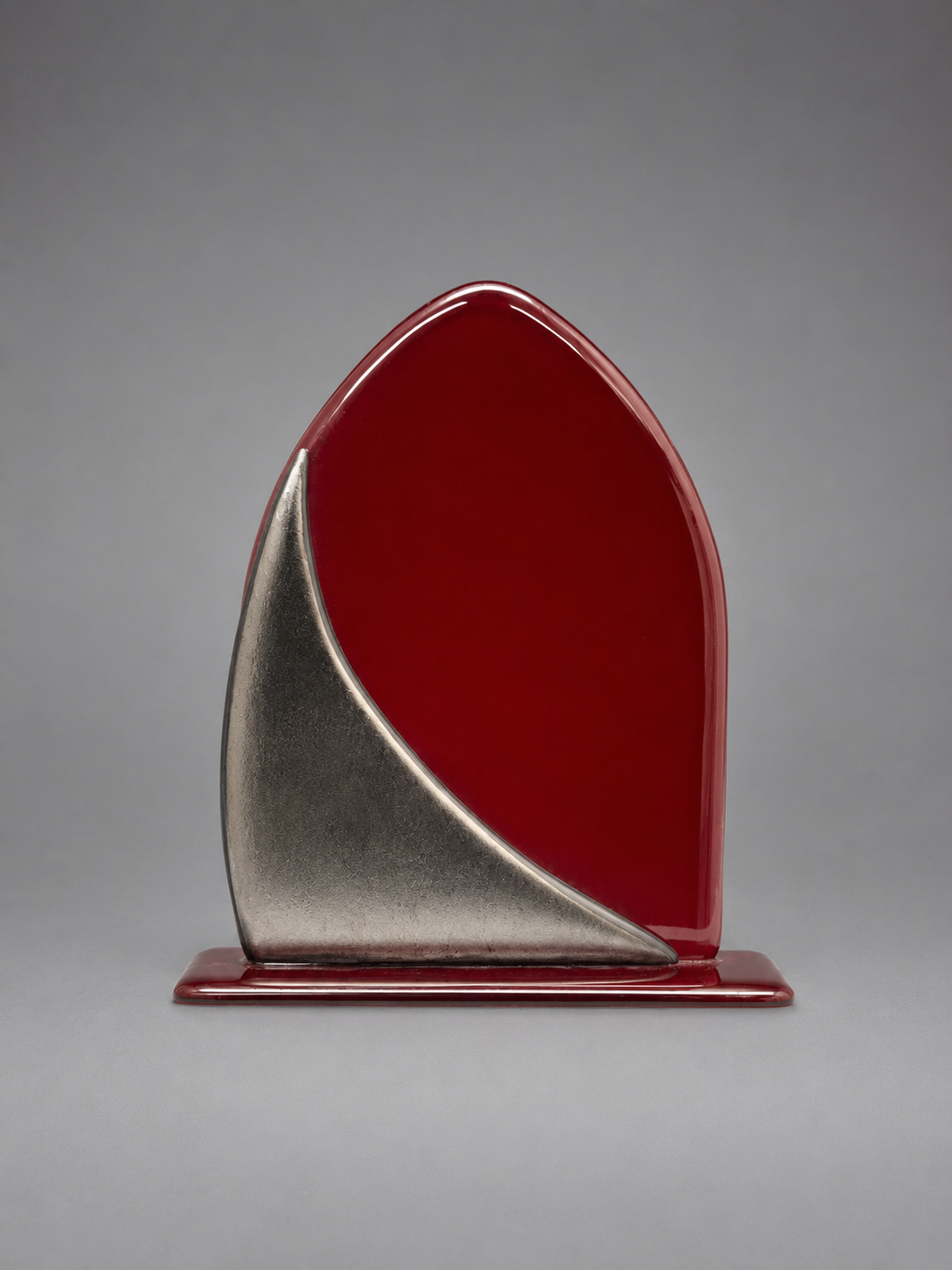 A modern abstract glass plaque featuring a glossy deep red arch shape with a curved metallic silver front panel and a matching red base, set against a neutral gray background.