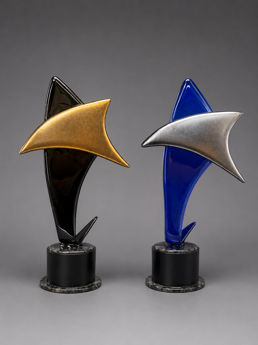 Two modern abstract trophy or sculpture pieces on round black bases, shown against a smooth gray studio background. The left piece has a black and gold design, and the right piece has a blue and silver design.