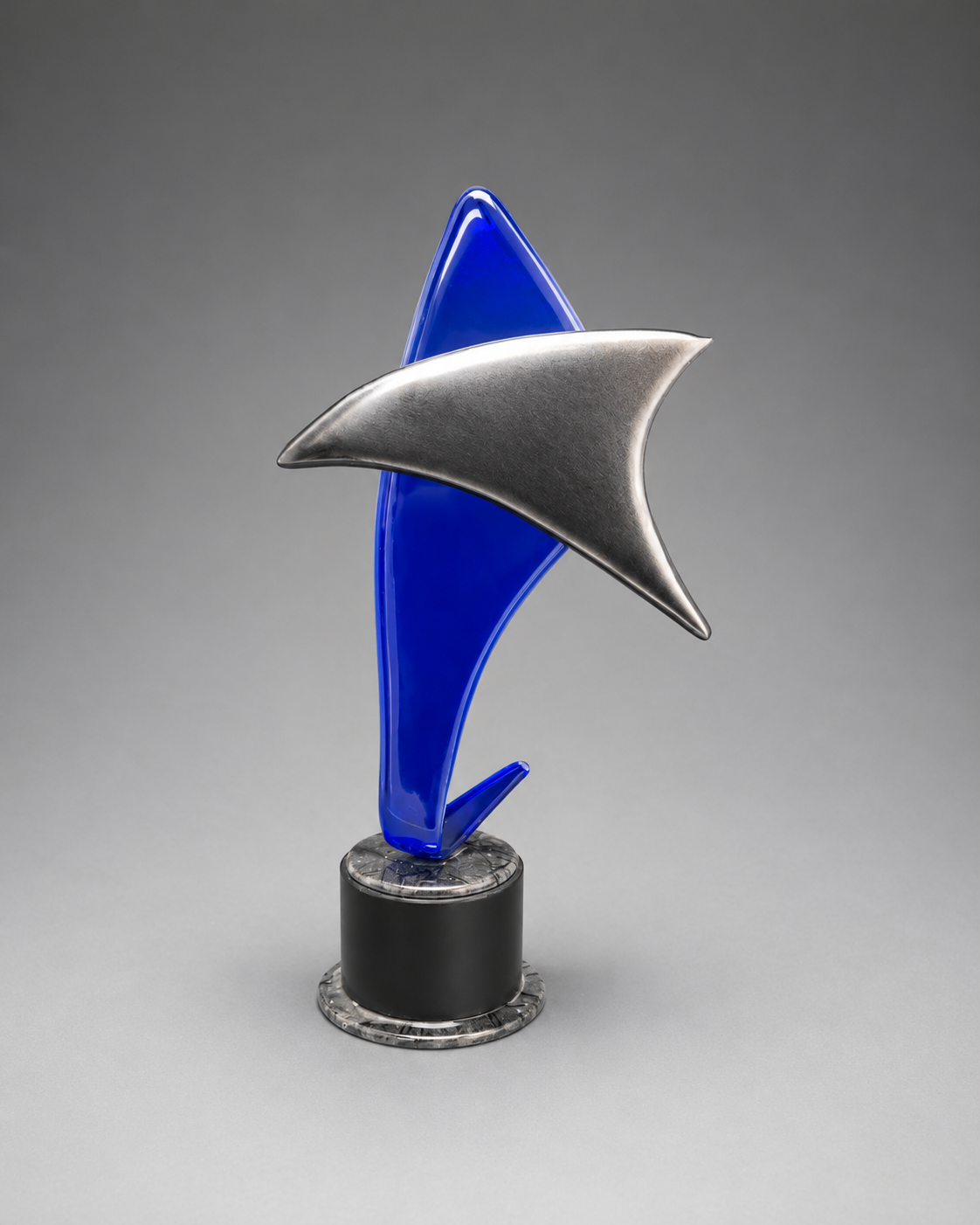 A modern abstract trophy or sculpture featuring a metallic silver delta-shaped fin layered over a glossy royal blue vertical form, mounted on a black cylindrical base with a silver circular rim, photographed on a neutral gray gradient background.