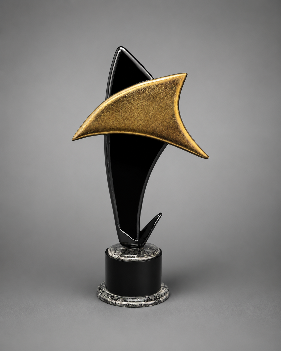 A modern abstract trophy sculpture featuring a black glossy curved base, a textured metallic gold delta-shaped top, and a round black and silver marble-patterned pedestal, set against a neutral gray studio background.