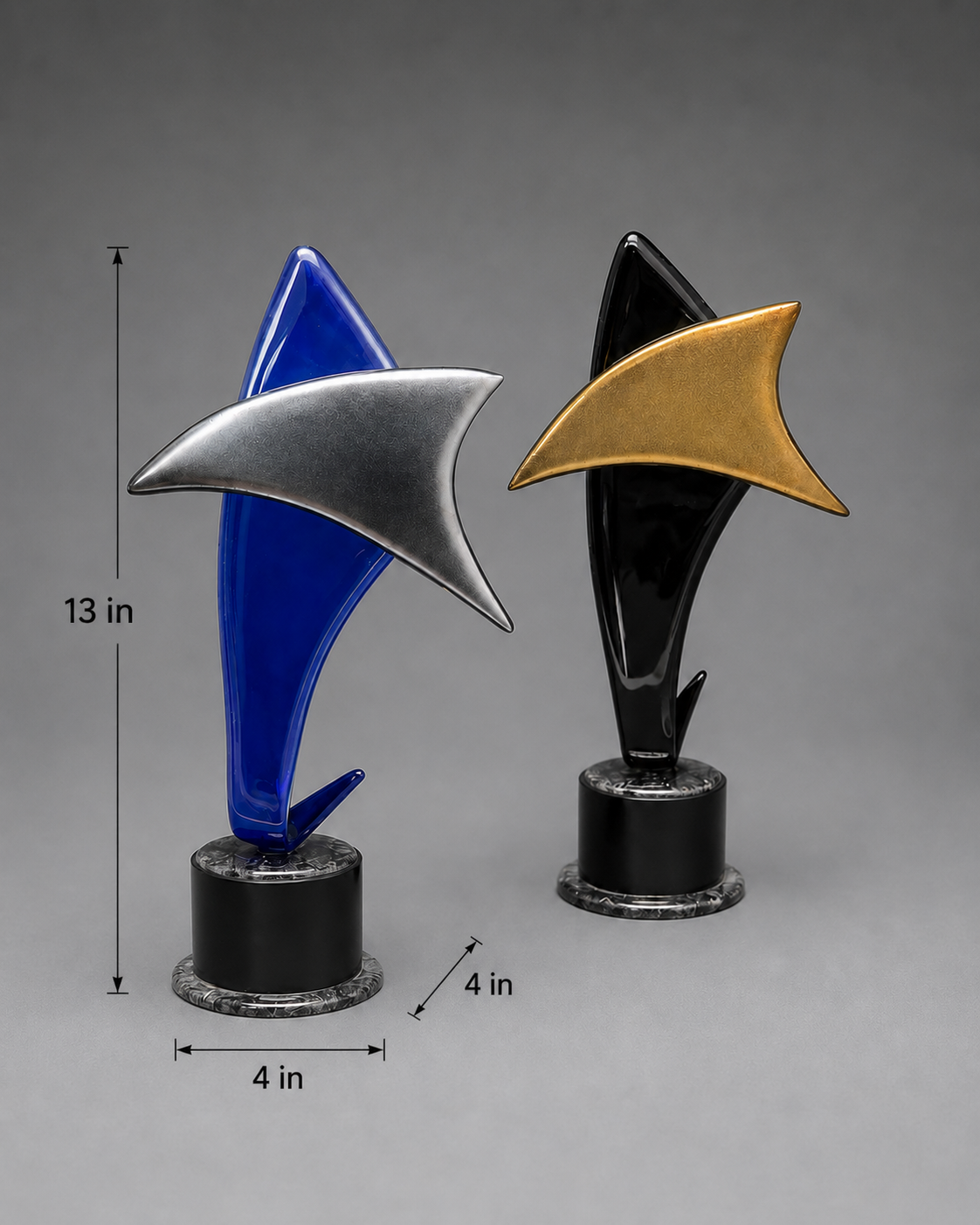 Two modern shark-shaped trophy or award sculptures on black cylindrical bases, shown against a smooth gray studio background. One sculpture is blue with a silver shark fin overlay, and the other is black with a gold shark fin overlay. Measurements: 4 inches width, 4 inches length, and 13 inches height.