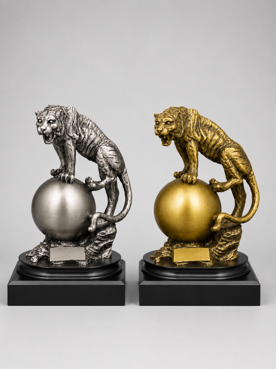 Two tiger figurines on spherical bases, one silver and one gold, on a gray background.