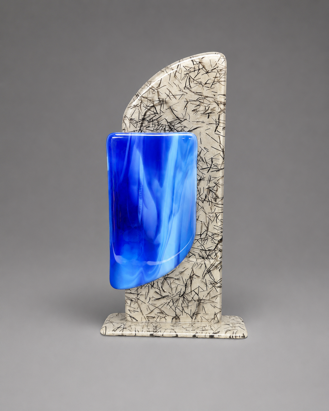 A sculptural glass plaque with a vibrant blue curved body and a cream-colored backplate decorated with black linear patterns, set against a neutral gray background.