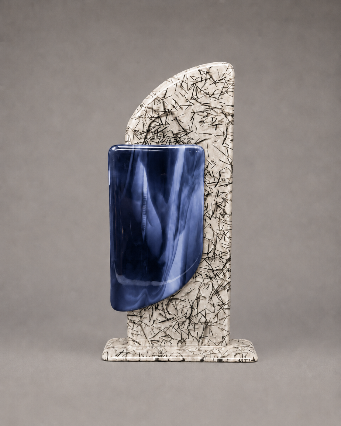 A modern sculptural glass plaque with a glossy deep blue curved front and a textured off-white back panel with black linear patterns, set against a neutral gray background.